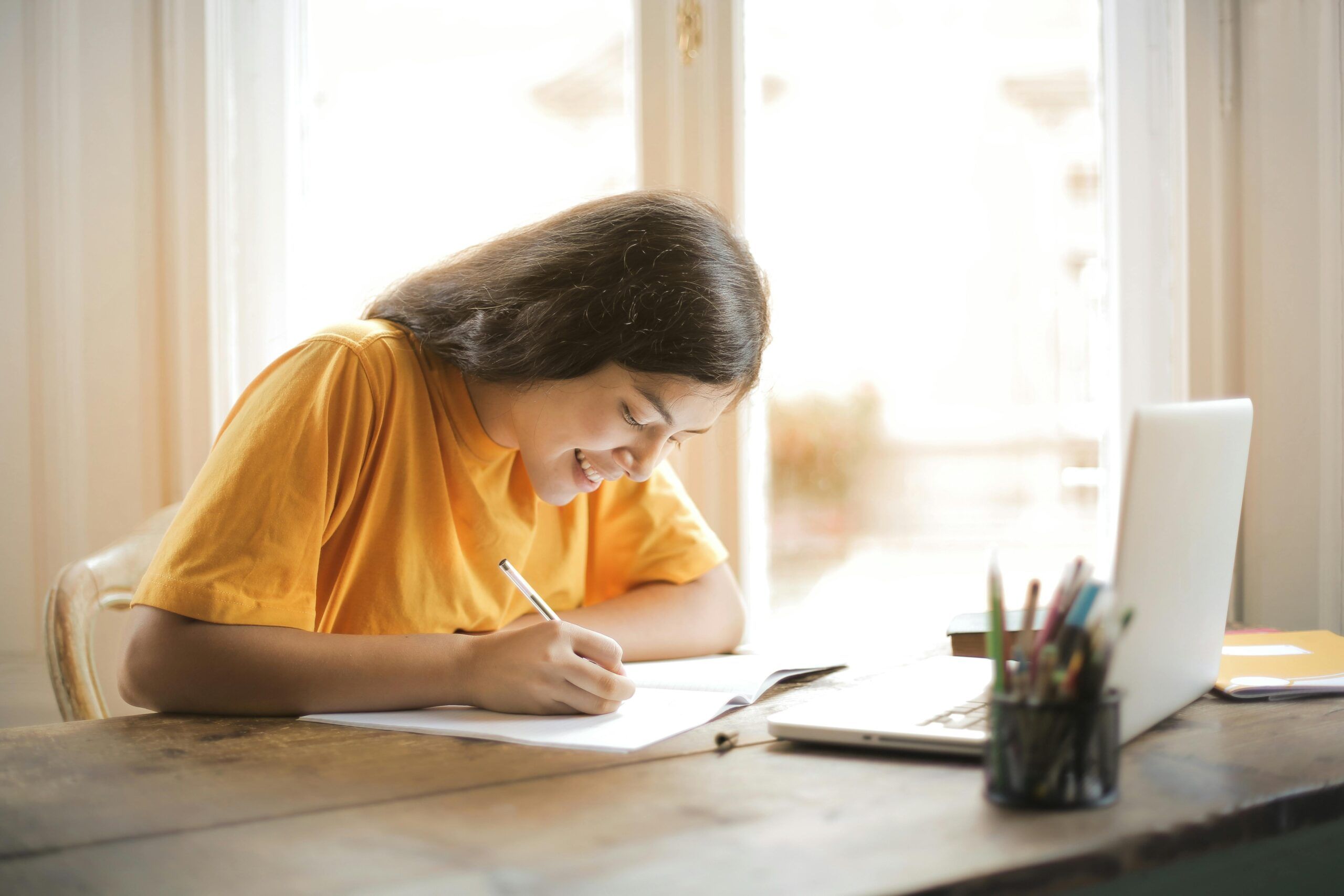 Photo of young woman sitting at a desk with a window behind her. She's writing in a notebook, with a computer open in front of her.