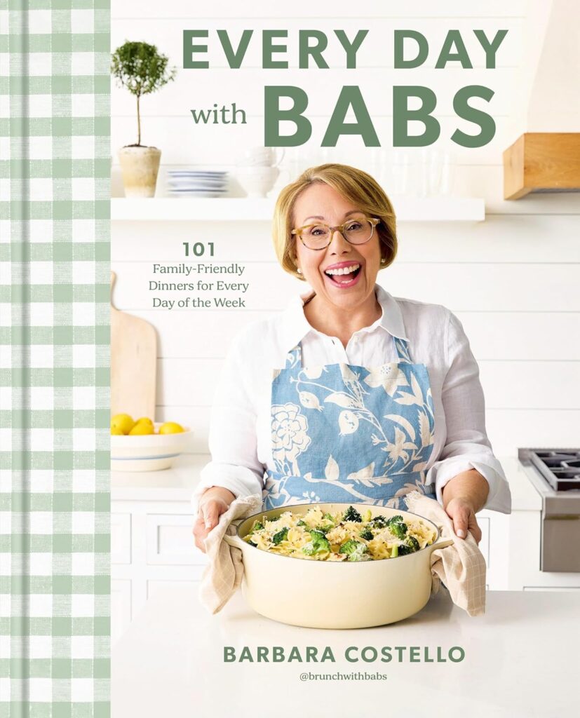 Cover of Every Day with Babs. Author Barbara Costello is standing in a white kitchen with open shelves behind her. She's wearing a white shirt and a blue floral apron, and has a pot of pasta with broccoli in her hands.