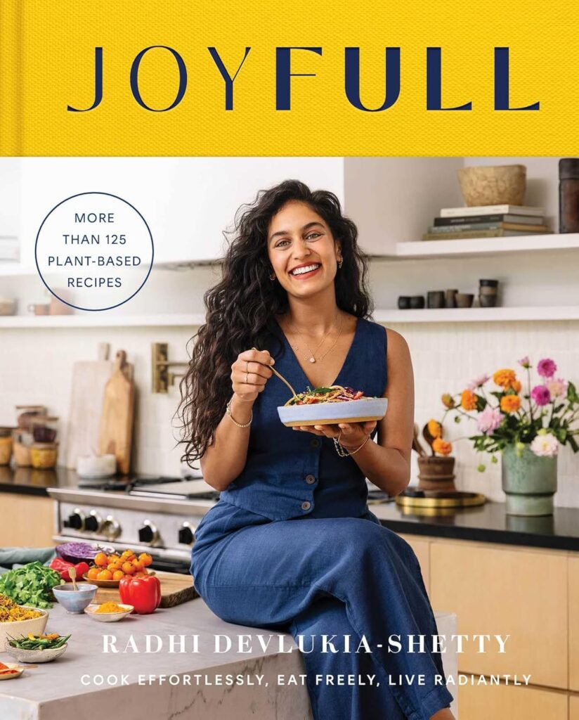 Cover of Joyfull. Author Radhi Devlukia-Shetty is sitting on her kitchen counter, wearing a blue vest and pants. She is holding a dish with vegetables and pasta like she's getting ready to eat it. There are vegetables in dishes on the counter to her right, and a kitchen with open shelves behind her.
