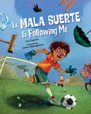 Cover of La Mala Suerte is Following Me. Latine boy looks afraid as he runs across a soccer field. There's a black umbrella, a broken mirror, a ladder, and a black bird above him, and a soccer ball with a scary shadow with eyes at his feet.