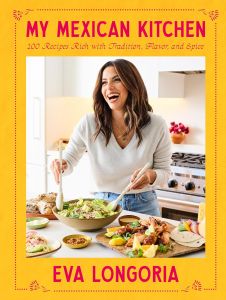 Cover of My Mexican Kitchen. Author Eva Longoria is standing in her kitchen, looking off-screen and laughing. She has serving spoons in her hands, like she's getting ready to place some of the salad in the bowl on a plate. There is more food in plates and bowls around her.